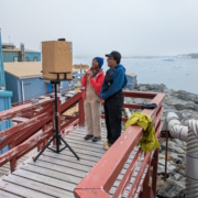 Two scientists on the deck of Palmer Station in Antarctica speaking to students over a laptop with glaciers and ocean behind them.