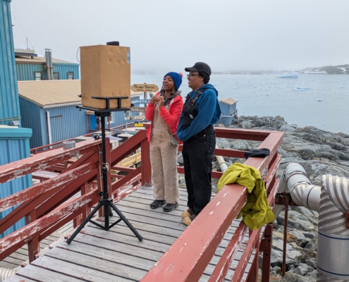 Two scientists on the deck of Palmer Station in Antarctica speaking to students over a laptop with glaciers and ocean behind them.