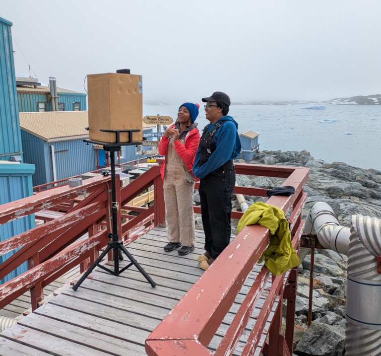 Two scientists on the deck of Palmer Station in Antarctica speaking to students over a laptop with glaciers and ocean behind them.