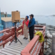 Two scientists on the deck of Palmer Station in Antarctica speaking to students over a laptop with glaciers and ocean behind them.