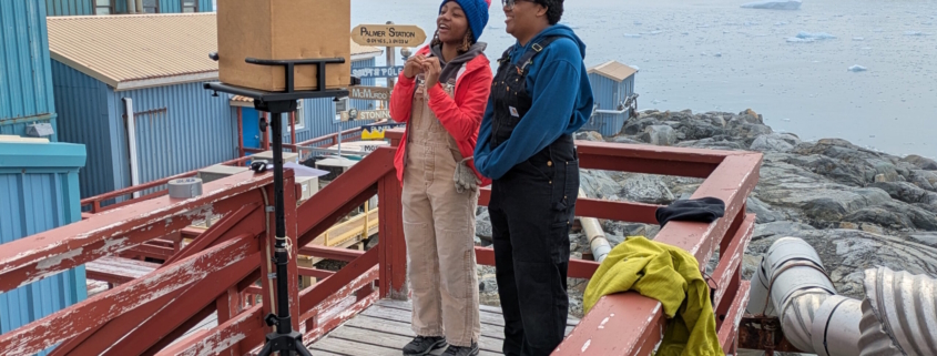 Two scientists on the deck of Palmer Station in Antarctica speaking to students over a laptop with glaciers and ocean behind them.