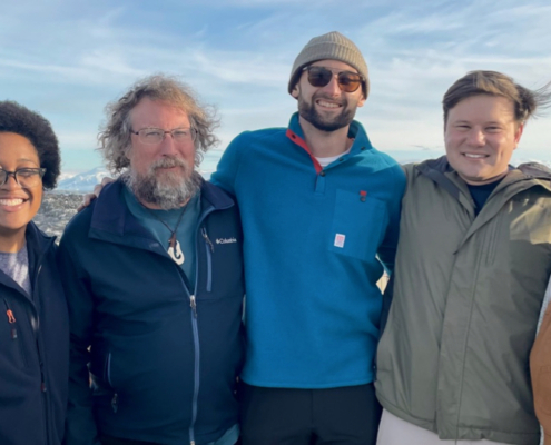 5 people standing on a rocky beach with mountains in the background.