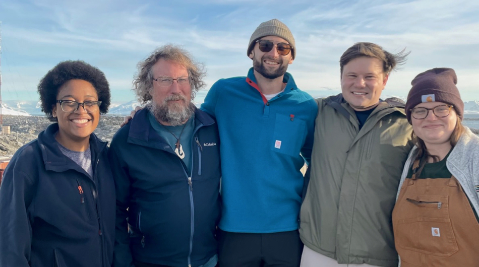 5 people standing on a rocky beach with mountains in the background.